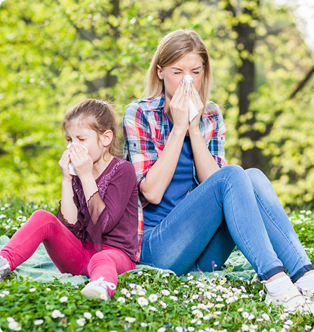 Mother and daughter sitting on a hill outside and blowing their noses, in need of an ENT for allergy treatment.