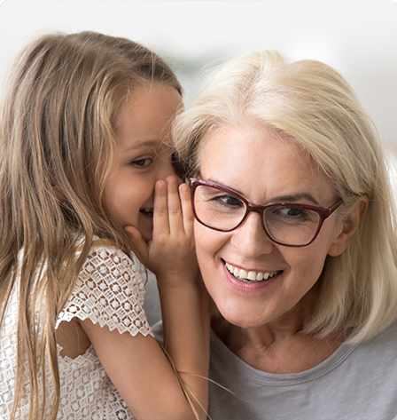Grandchild whispering into her grandmother's ear, after her grandmother got an evaluation at her local hearing and balance clinic.