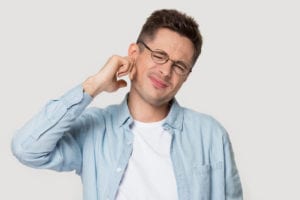 Studio head shot portrait on grey background frowning young man in glasses touch ear closed it with finger hurt suffers from earache strong pain, guy look at camera annoyed by loud noise concept image