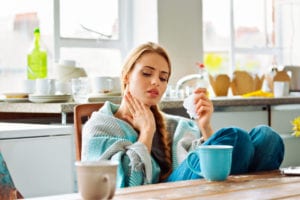 Young woman has a cold, sitting in a kitchen wrapped in blanket and touching her neck.