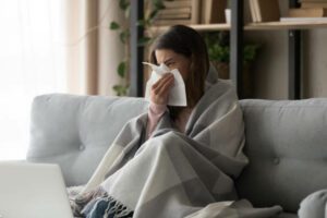 Sick young woman sitting on couch at home, unhealthy girl covered with warm plaid sneezing blowing her nose on paper tissue suffering from grippe temperature or seasonal allergy, cold dwelling concept