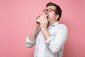 Young caucasian man with handkerchief and glasses sneezing being ill or having allergy. Studio shot on pink wall.