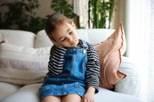 Young girl, in need of ear infection treatment, sitting on a couch and holding her ear in discomfort.