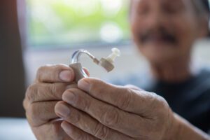 A blurry image of a smiling senior man with hearing loss is holding a hearing aid at home, looking at it with satisfaction.