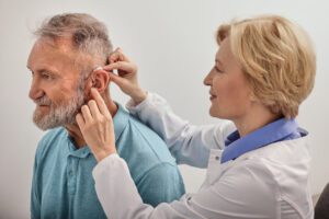 Experienced female doctor fitting Behind-The-Ear hearing aid on senior patient's ear while consultation in audiology clinic